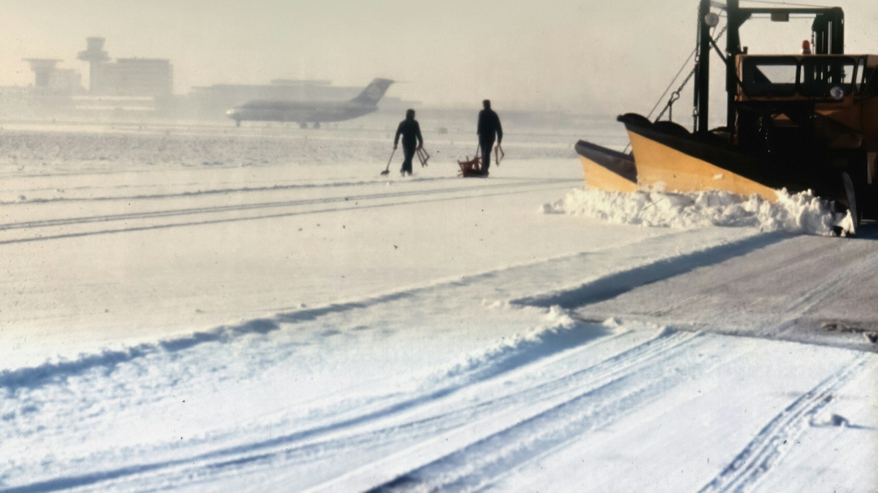 Two airport workers clearing snow from an apron beside a snow plough blade